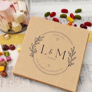 May include: A square brown paper bag featuring a black circular wreath design with the letters L & M, the date 29.08.2026, and the text THANK YOU FOR SHARING OUR SPECIAL DAY with two small heart icons printed in the centre of the design.