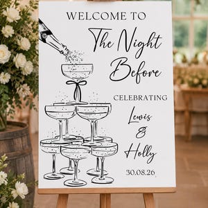 May include: A white rectangular sign on a wooden easel featuring black line art of a champagne bottle pouring into a stacked tower of coupe glasses. The text reads: WELCOME TO The Night Before CELEBRATING Lewis & Holly 30.08.26 in black font.