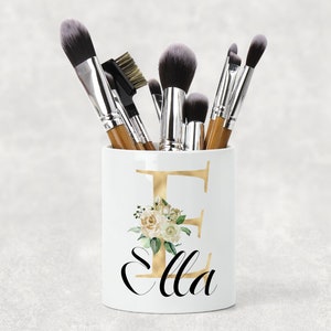 May include: White ceramic makeup brush holder with a gold letter "E" and the name "Ella" in black script. The holder is filled with various makeup brushes.