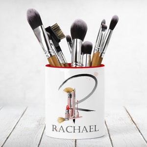 May include: White ceramic makeup brush holder with a red rim. The holder features a black and white graphic of the letter "R" with makeup brushes and lipstick. The text "RACHAEL" is printed below the graphic.
