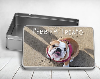 May include: A rectangular metal tin with an open lid. The tin's surface features a photo of a bulldog wearing a pink harness, with the text "PEBBLES TREATS" in white. The dog is on a sandy surface.