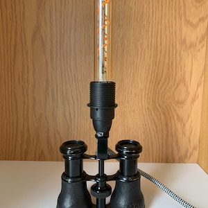 Peut inclure: Une lampe de table unique avec une base de jumelles d'opéra noires vintage. La lampe a une ampoule claire et allongée avec un filament orange. La lampe est branchée sur une prise murale avec un cordon rayé noir et blanc.
