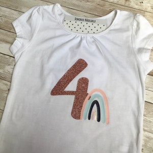 May include: White t-shirt featuring a copper glitter number 4 and a pastel rainbow design. The top has a round neck and short sleeves. The text "SCHICKES BÜGELBILD" is printed on a tag inside the collar.