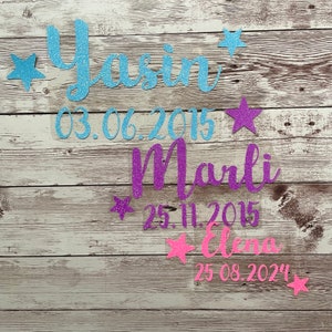 Könnte beinhalten: Drei Namen mit Geburtsdaten in Glitzer auf einem Holzgrund. Die Namen sind Yasin 03.06.2015, Marli 25.11.2015 und Elena 25.08.2024. Jeder Name hat einen Stern daneben.