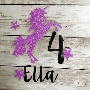 Könnte beinhalten: Ein lila Glitzer-Einhorn-Kuchenaufsatz mit der Zahl 4 und dem Namen "Ella" in schwarzer Schrift. Der Aufsatz enthält lila Glitzersterne. Das Design befindet sich auf einer transparenten Rückseite.