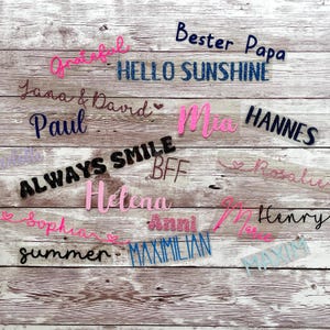 May include: A collection of iron-on patches with various designs and text, including "Bester Papa", "Hello Sunshine", "Grateful", "Jana & David", "Paul", "Always Smile", "Mia", "Hannes", "BFF", "Helena", "Anni", "Henry", "Sophia", "Summer", and "Maximilian".