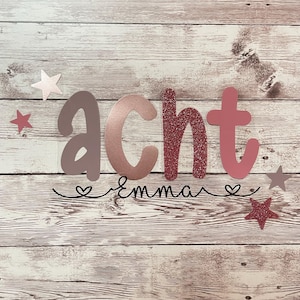 Peut inclure: Une décoration murale en lettres scintillantes rose, or rose et marron qui dit "acht" avec le nom "emma" écrit en cursive en dessous. La décoration murale est sur un fond en bois blanc avec de petites étoiles roses et dorées.