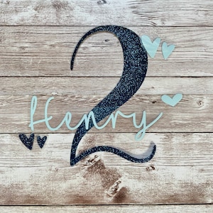 Könnte beinhalten: Eine blaue Glitzerzahl Zwei mit dem Namen Henry in weißem Glitzer, umgeben von kleinen weißen Glitzerherzen auf einem Holzgrund.
