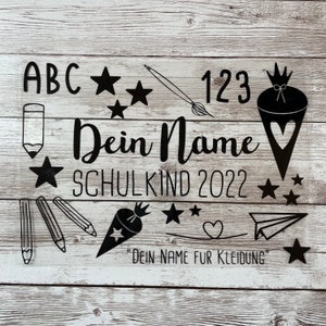Könnte beinhalten: Schwarz-weißes Bügelbild für Kleidung. Das Design beinhaltet die Buchstaben ABC, die Zahlen 123, Sterne, Bleistifte, einen Pinsel, ein Herz, eine Karotte und ein Papierflugzeug. Der Text "Dein Name" und "Schulkind 2022" sind ebenfalls im Design enthalten. Der Text "Dein Name fur Kleidung" befindet sich am unteren Rand des Designs.