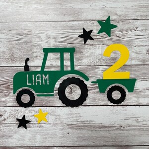 Puede incluir: Un tractor verde con un número 2 amarillo en la parte trasera. El tractor tiene el nombre Liam escrito en el lateral. Hay cinco estrellas en el fondo, dos negras y tres verdes.
