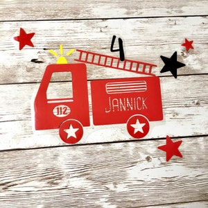 May include: Red fire engine with a ladder, a yellow light, and the number 112 on the side. The engine has the name "JANNICK" written on the side. There are five red stars and one black star on a wood background. The number 4 is written in black above the engine.