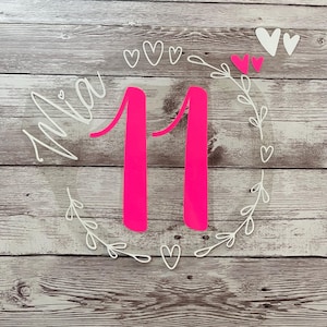 Könnte beinhalten: Ein weißer Acrylkreis mit einer pinkfarbenen Zahl 11, dem Wort "Mia" in weißer Schrift und einem weißen Kranz mit weißen Herzen um die Zahl.