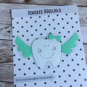 Könnte beinhalten: Ein weißer Bügelbild-Transfer einer Zahn mit Flügeln und einer Schleife. Der Transfer befindet sich auf einem weißen Hintergrund mit schwarzen Sternen. Der Text "Schickes Bügelbild" und "Für alle hellen oder dunklen Textilien geeignet." ist auf dem Transfer gedruckt.