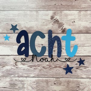 Könnte beinhalten: Blaue, graue und hellblaue Filzbuchstaben, die "acht" buchstabieren, mit einem schwarzen kursiv geschriebenen Namen "Noah" und blauen Glitzersternen auf einem weißen Holzgrund.