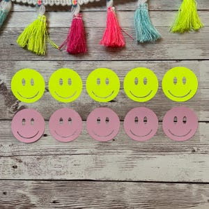 Op de afbeelding: Tien smiley-stickers, vijf zijn neon geel en vijf zijn roze. De stickers zijn gerangschikt in twee rijen van vijf op een houten oppervlak.
