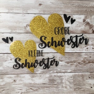 Könnte beinhalten: Zwei goldene Glitzerherzen mit schwarzem Glitzertext. Das größere Herz sagt "GROBE SCHWESTER" und das kleinere Herz sagt "KLEINE SCHWESTER".