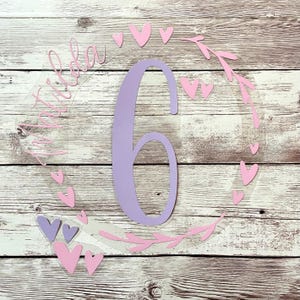 Puede incluir: Un círculo acrílico transparente con un número seis morado y el texto "Ava tiene seis años" en letra cursiva. El círculo está decorado con corazones rosas y hojas.