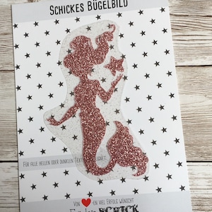 Peut inclure: Un patch thermocollant en forme de silhouette de sirène rose pailletée avec une étoile. Le patch est sur un fond blanc avec un motif d'étoiles noires. "Schickes Bügelbild" et "Für alle hellen oder dunklen Textilien geeignet." sont écrits en texte noir.  "Von Herzen viel Erfolg wünscht Euch ein Schick" est écrit en texte noir avec un cœur rouge.