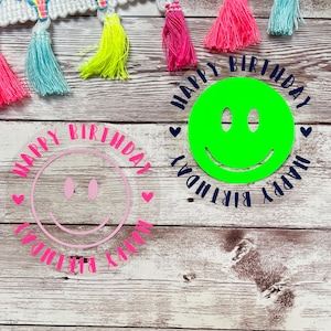 Peut inclure: Deux décorations rondes en acrylique pour gâteau avec un motif de visage souriant. Une décoration est rose et dit "Joyeux anniversaire" en blanc. L'autre décoration est verte et dit "Joyeux anniversaire" en bleu marine.