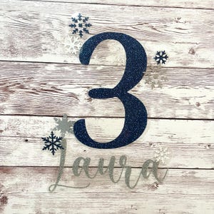 Peut inclure: Un chiffre 3 bleu marine pailleté avec le nom "Laura" en script argenté pailleté, entouré de flocons de neige argentés et bleu marine. Le design est posé sur un fond en bois clair rustique.