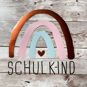 Könnte beinhalten: Ein Regenbogen-Aufkleber mit einem kupferfarbenen, rosa und blauen Design. Der Regenbogen hat ein kleines Herz in der Mitte. Der Text "SCHULKIND" befindet sich unter dem Regenbogen.