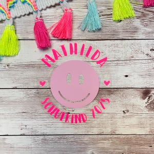 Peut inclure: Un autocollant circulaire rose et blanc avec le texte "MATHILDA" et "SCHOOL KIND 2023" autour d'un smiley rose.