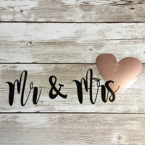 Könnte beinhalten: Ein transparenter Aufkleber mit dem Text "Mr & Mrs" in schwarzer Schrift. Ein roségoldenes Herz ist über dem Text platziert.