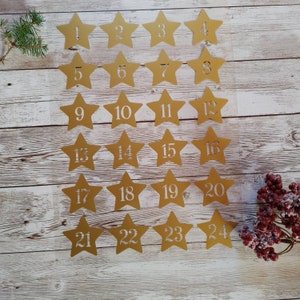 May include: Twenty-four gold star-shaped paper ornaments with white numbers from 1 to 24. The ornaments are arranged in four rows of six on a white wooden background.
