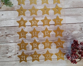 Könnte beinhalten: Vierundzwanzig goldene sternförmige Papierschnitte mit den Zahlen 1 bis 24 darauf gedruckt. Die Sterne sind in vier Reihen zu je sechs Sternen auf einem hellbraunen Holzhintergrund angeordnet.