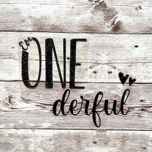 Pode incluir: Um letreiro preto com glitter "One derful" com dois pequenos corações pretos. O letreiro é feito de madeira e tem um visual rústico.
