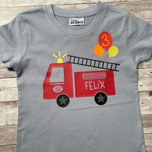 Könnte beinhalten: Ein graues T-Shirt mit einem roten Feuerwehrauto-Motiv. Das Auto hat eine Leiter, ein gelbes Licht und die Nummer 112 an der Seite. Die Worte "Felix" und "3" stehen ebenfalls auf dem Shirt.