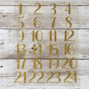 Può includere: Un foglio di numeri dorati glitterati su uno sfondo trasparente. I numeri sono da 1 a 24.