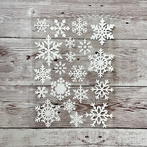 Könnte beinhalten: Eine Sammlung weißer Papierschneeflocken in verschiedenen Größen, angeordnet auf einem hellen Holzhintergrund.