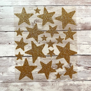 May include: A sheet of gold glitter star stickers. The stars are various sizes and are arranged in a random pattern.