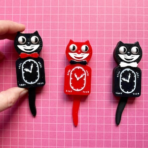 May include: Three novelty cat-shaped clocks in black and red, each with a bow tie and a clock face. One black cat clock is held by a hand. The clocks have a retro design and are set against a pink grid background.