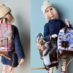 以下が含まれることがあります： ニット帽と紺色のセーターを着た人形が、いくつかのミニチュアバックパックを持っています。バックパックは、花柄、パッチワーク、人魚のデザインなど、さまざまなパターンが特徴です。各バックパックには、茶色の革のアクセントと小さなフロントポケットが付いています。