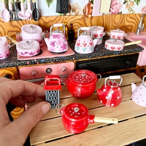 Może przedstawiać: Miniaturowy zestaw kuchenny z różowymi i czerwonymi naczyniami. Zawiera garnki, czajniki i tarkę. Niektóre elementy mają wzory kwiatowe lub w kropki. Zestaw jest ułożony na drewnianej powierzchni.