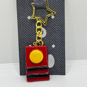 May include: A red and yellow charm with a gold chain and star charm. The charm is shaped like a rectangle with a yellow circle in the center and two black stripes.