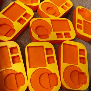 May include: A collection of bright orange and yellow 3D-printed objects. Each rectangular piece has a circular indentation, a square section, and two smaller square cutouts. The objects are arranged on a neutral-colored surface.