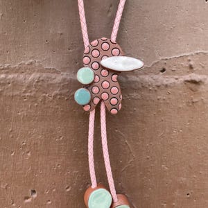 May include: A bolo tie featuring a pink braided cord and a central ceramic pendant with a brown and pink polka dot pattern, accented by two small light green circular beads and a white horizontal oval shape, ending in two light green bead tips.
