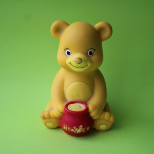 Könnte beinhalten: Ein gelber Gummibär, der einen roten Honigtopf mit dem Wort "Honey" hält. Der Bär hat ein freundliches Gesicht und sitzt auf grünem Hintergrund.