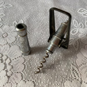 May include: A silver corkscrew with a black metal lever and a silver handle. The corkscrew is on a white lace tablecloth.