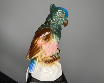 May include: A ceramic parrot figurine with a glossy finish. The bird features a teal head, brown and pink wings, and a white base. The parrot is perched on a branch, showcasing a detailed and colorful design.