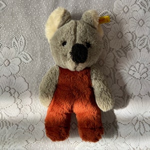Op de afbeelding: Een grijze koala teddybeer met een bruine jumpsuit. De beer heeft een zwarte neus en ogen. De beer zit op een witte kant tafelkleed.