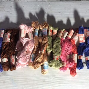 May include: A collection of 12 spools of embroidery floss in various colors, including pink, brown, blue, green, and red. The spools are wrapped in white paper with red and blue lettering.