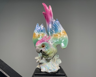 May include: A ceramic parrot figurine with outstretched wings, featuring a vibrant color palette. The bird's plumage showcases hues of pink, blue, green, and yellow. The figurine is mounted on a black base, creating a decorative accent.