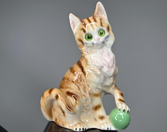 May include: A ceramic figurine of a tabby cat sitting with a green ball in its paws. The cat has green eyes and a white chest.