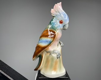 May include: A ceramic parrot figurine with a detailed design. The bird has a crest of pink feathers, a blue head, and a brown and blue wing. It is perched on a tree stump base with a white and tan color scheme. The parrot has an orange eye.
