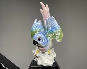 May include: A porcelain figurine of a blue and green parrot with pink accents. The parrot is perched on a white base with a black stand.