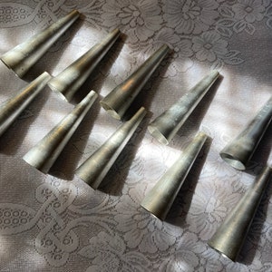 May include: A collection of ten silver-colored, cone-shaped metal molds. The molds are arranged on a white lace tablecloth, with sunlight casting shadows across the surface. The molds have a slightly aged appearance.